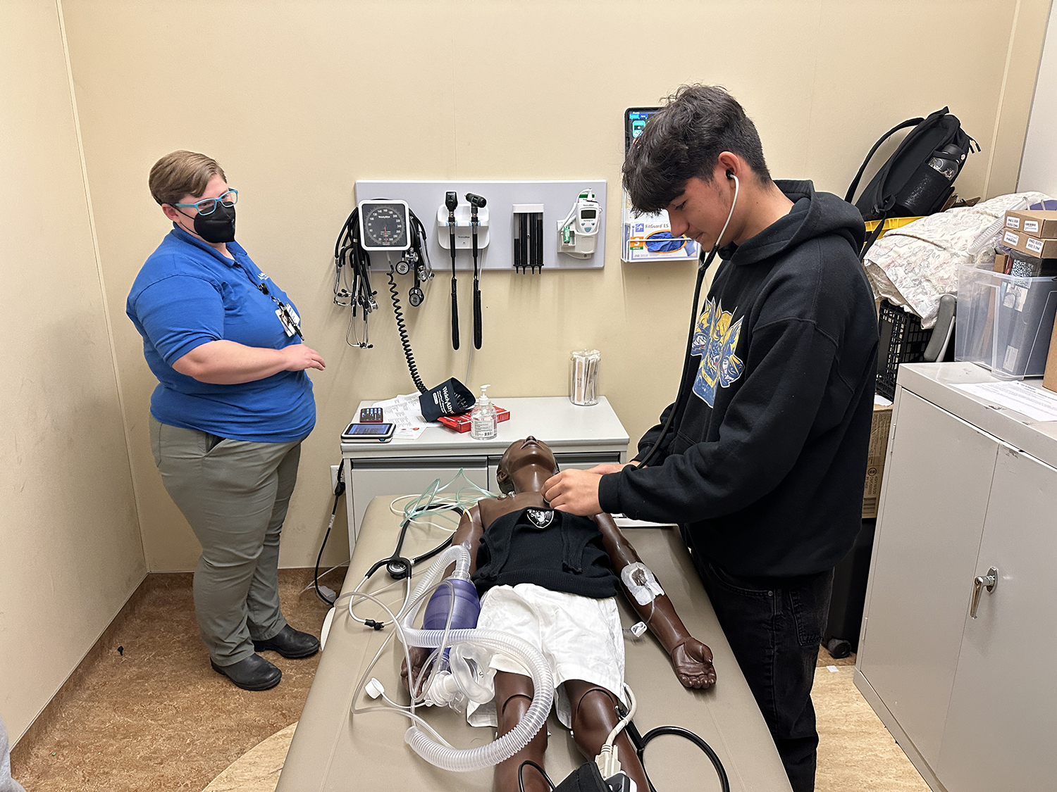 A high school student at a medical internship models using a stethoscope on a mannequin lying on a table in an exam room. A healthcare professional observes.