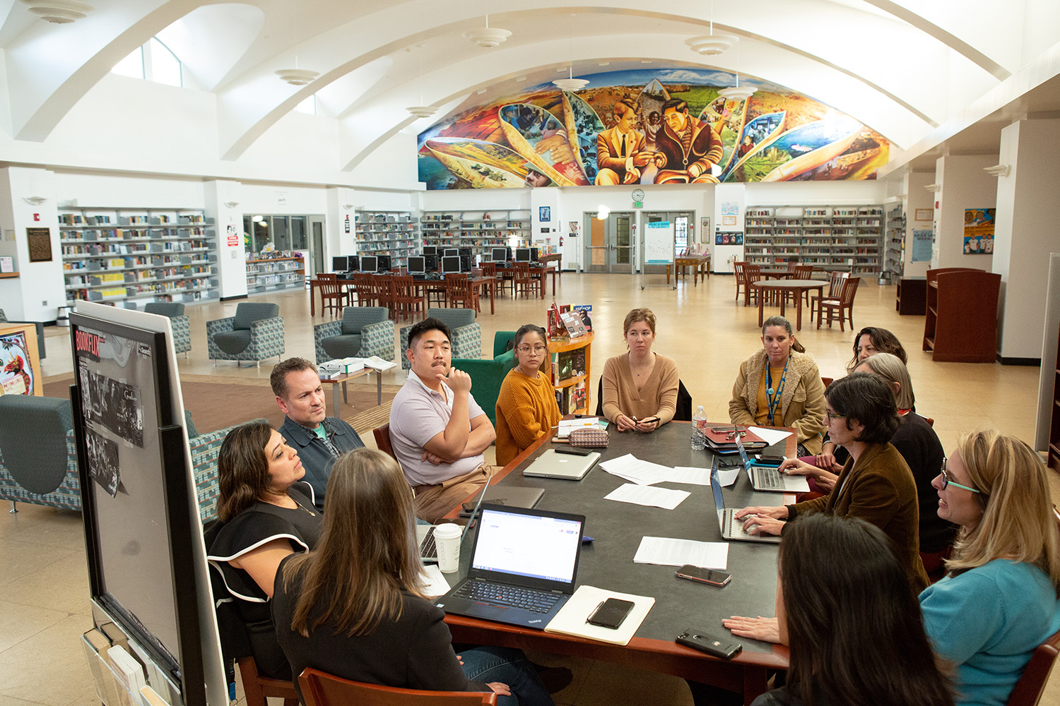 A group of teachers sit around a table with laptops and papers in a large school library.