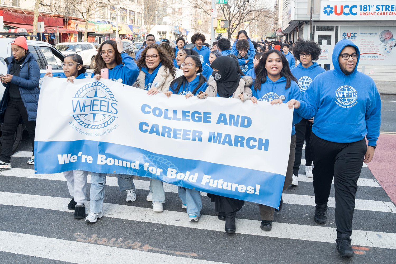 A large group of high school students wearing matching blue school sweatshirts walks across a city street, carrying a banner that says “WHEELS College and Career March - We’re bound for bold futures!”