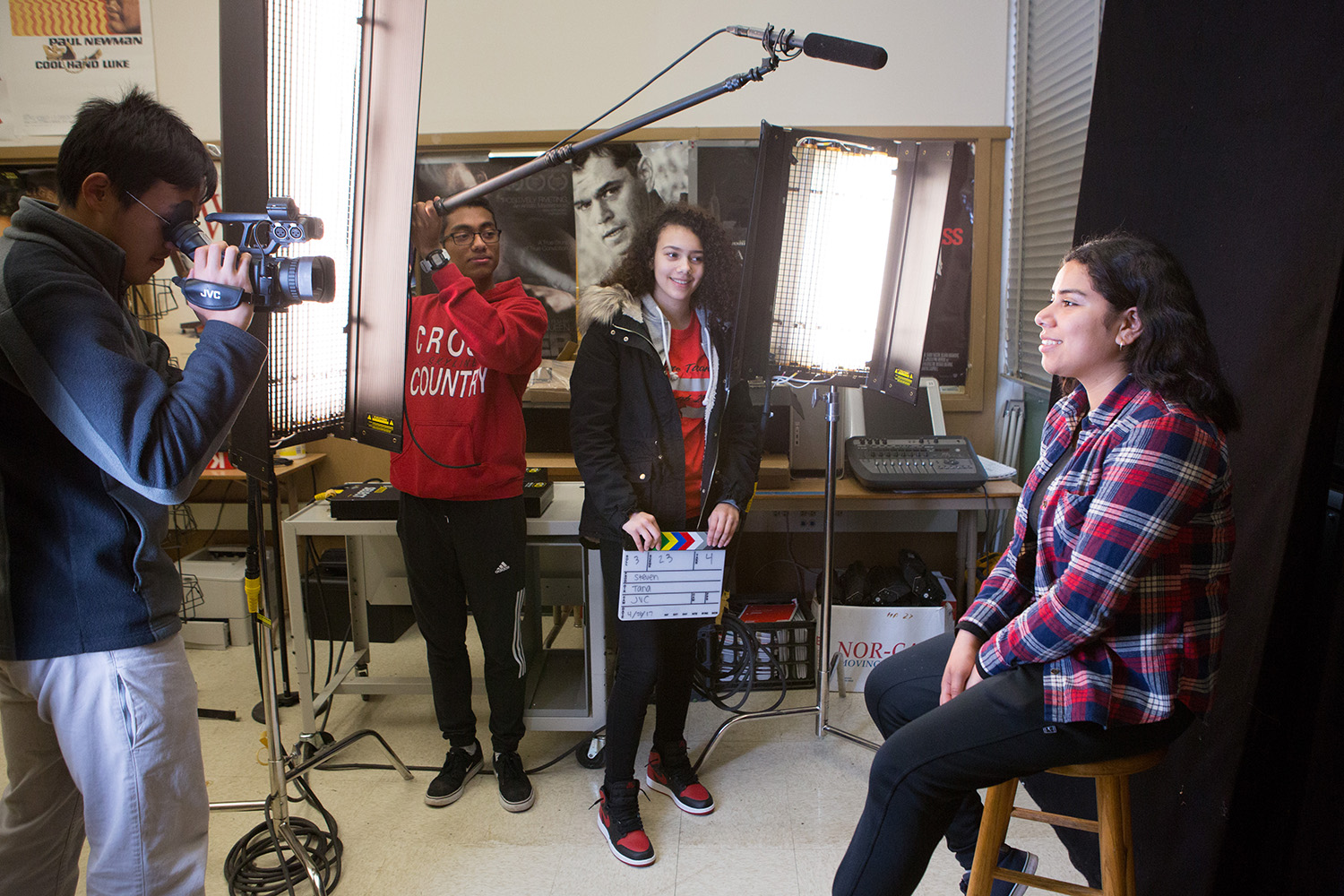 High school students work on a media project together: one holds a video camera while another holds a boom microphone, both pointing at a classmate sitting in front of a dark backdrop while another holds a film slate board to start a take.